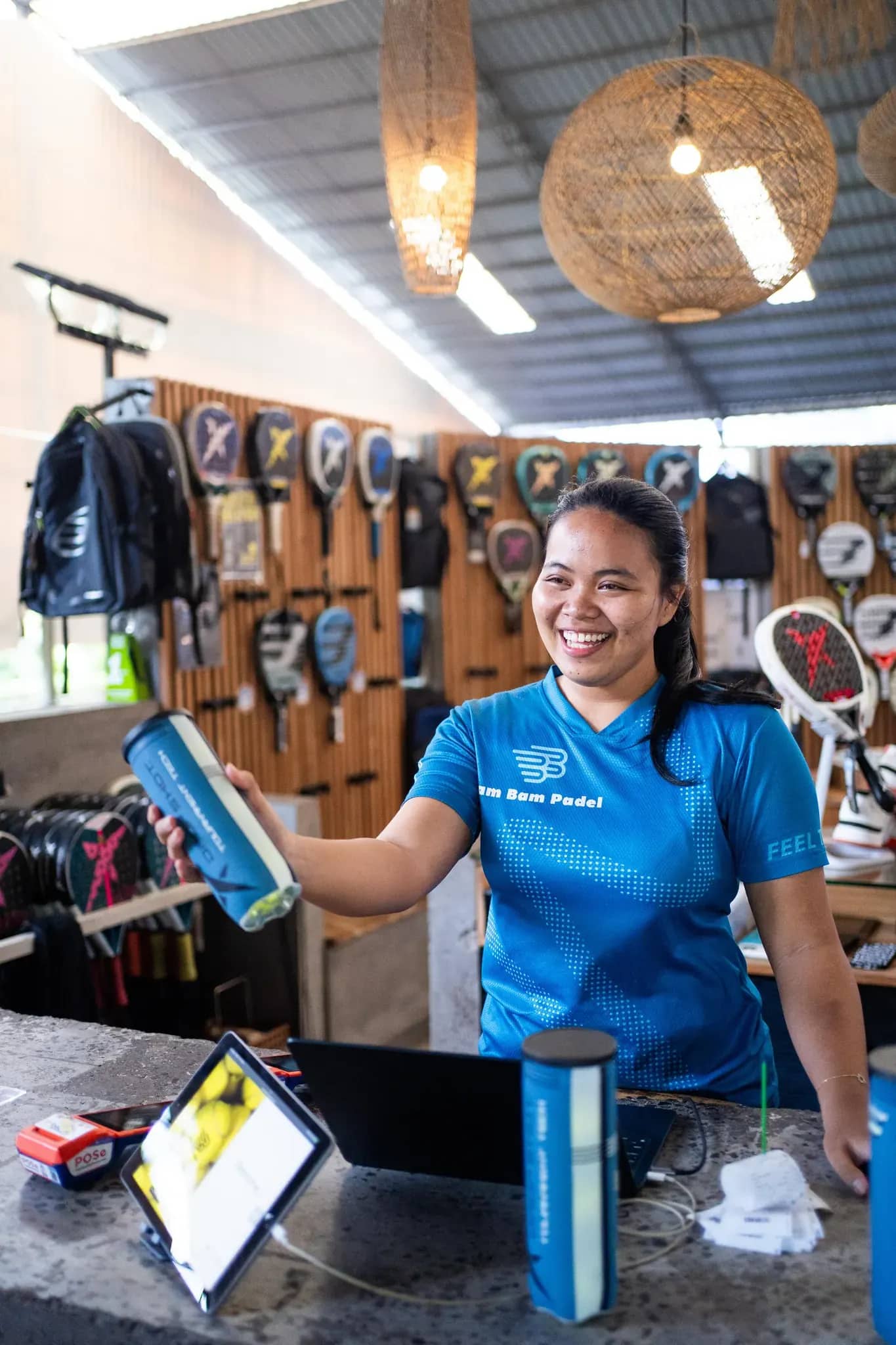 A pro-shop team member smiling at the counter, with the racket wall behind